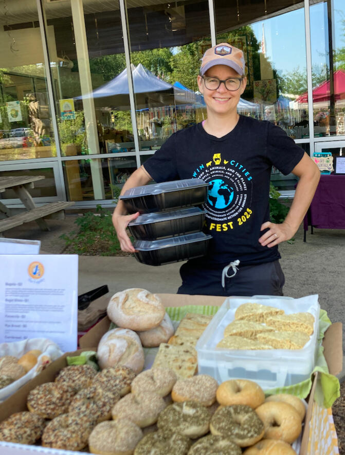 Photo of J. standing behind a farmers' market table of bagels, rye bread, focaccia, and cornbread. J. holds three tupperwares of additional baked goods.